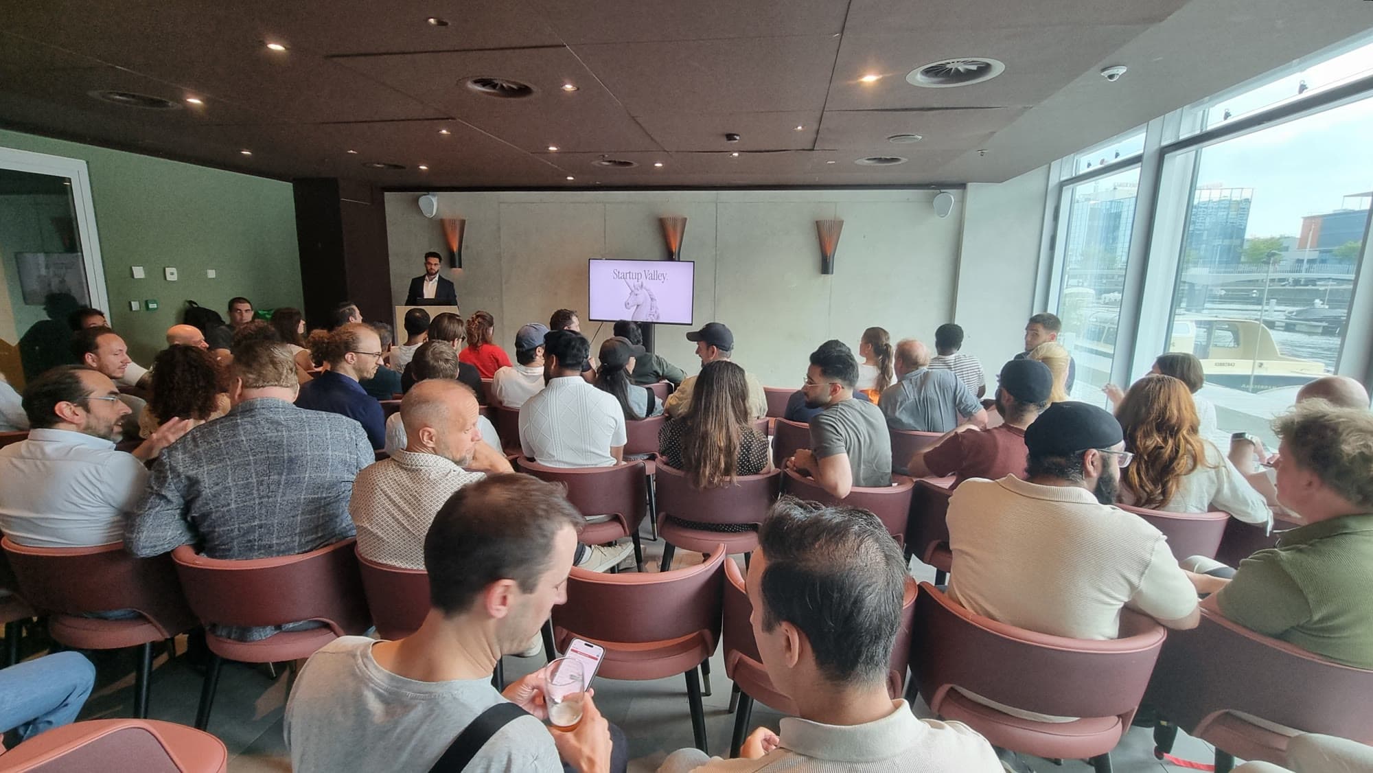 The full room at a Startup Valley Amsterdam pitch night at I Dock, harbour visible through the windows