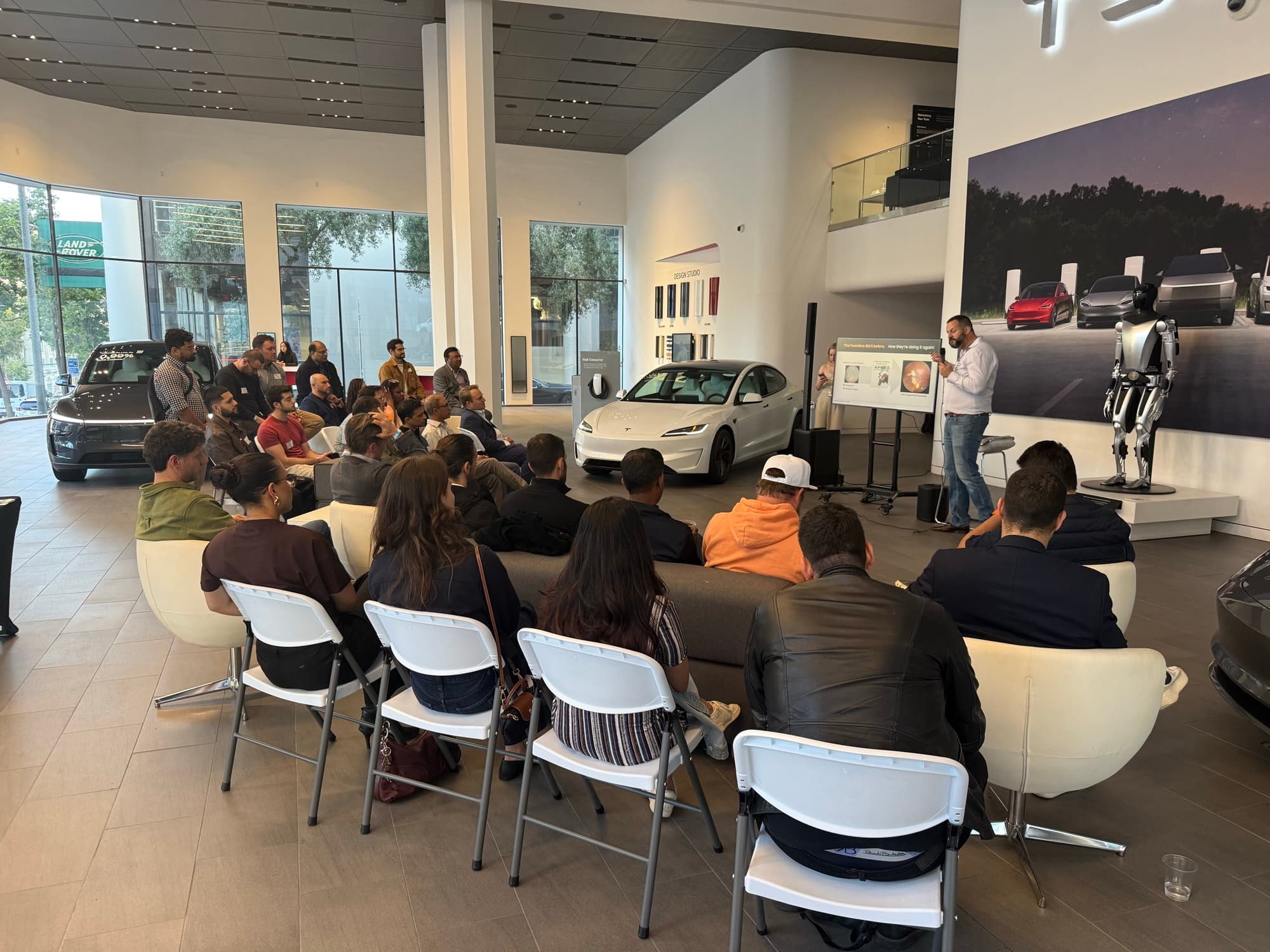 A founder pitching at a Startup Valley night in the Tesla showroom San Francisco, Optimus robot beside the stage